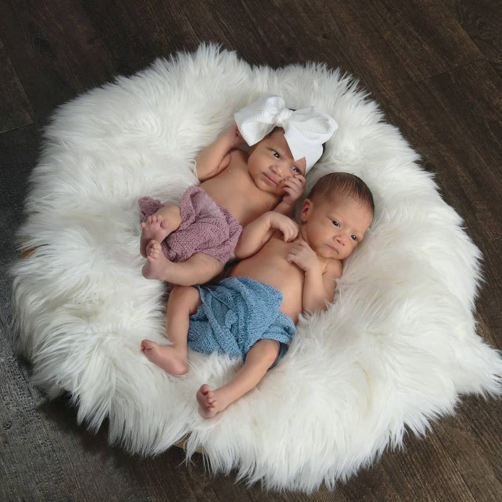 Newborn twins curled up together on a white fur blanket